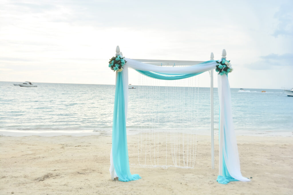 planning your dream wedding at sandals negril white angular arc with white and teal sheer fabric draped over it and crystals in the back positioned on the beach shore with small boats in the distance on a bright sunny day