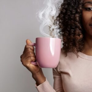 Black woman with curly auburn hair holding a pink mug with steam rising, smiling warmly.