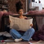 Illustrated fashion doll Julissa sitting on the floor of a sewing studio holding open a large sewing pattern with a confused expression — from the Introvert Stylist Julissa Collection