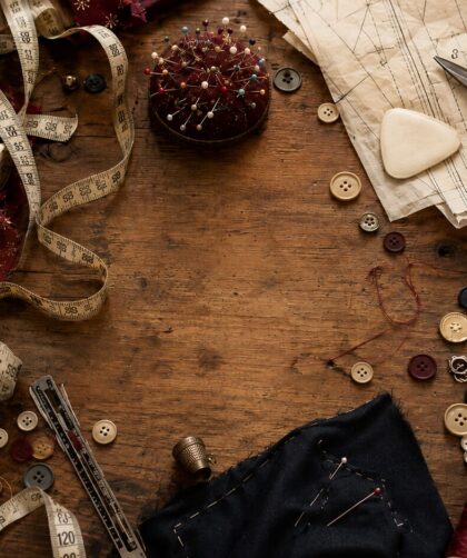 A flat lay of sewing notions scattered across a worn wooden table — thread spools, buttons, scissors, measuring tape, pin cushion, and pattern tissue paper — with one conspicuous empty space where the seam ripper should be.
